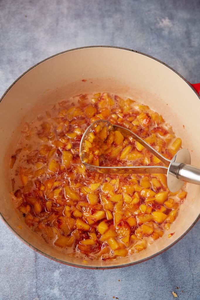 A potato masher smashing peaches in a pot.