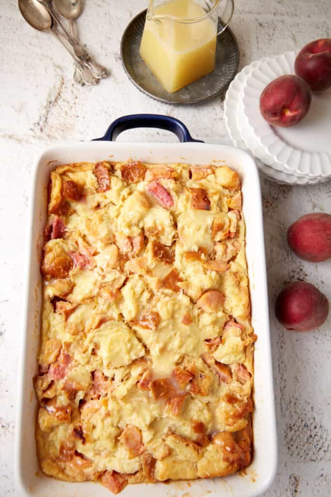 A baked peach bread pudding with a glaze in a pitcher next to it. 