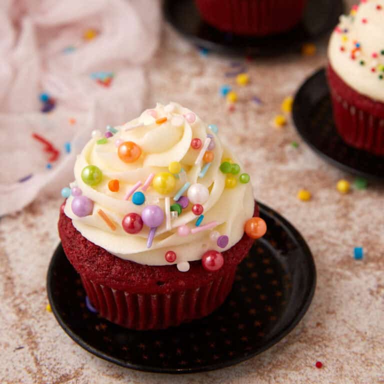 A single red velvet cupcake with cream cheese frosting covered in sprinkles.