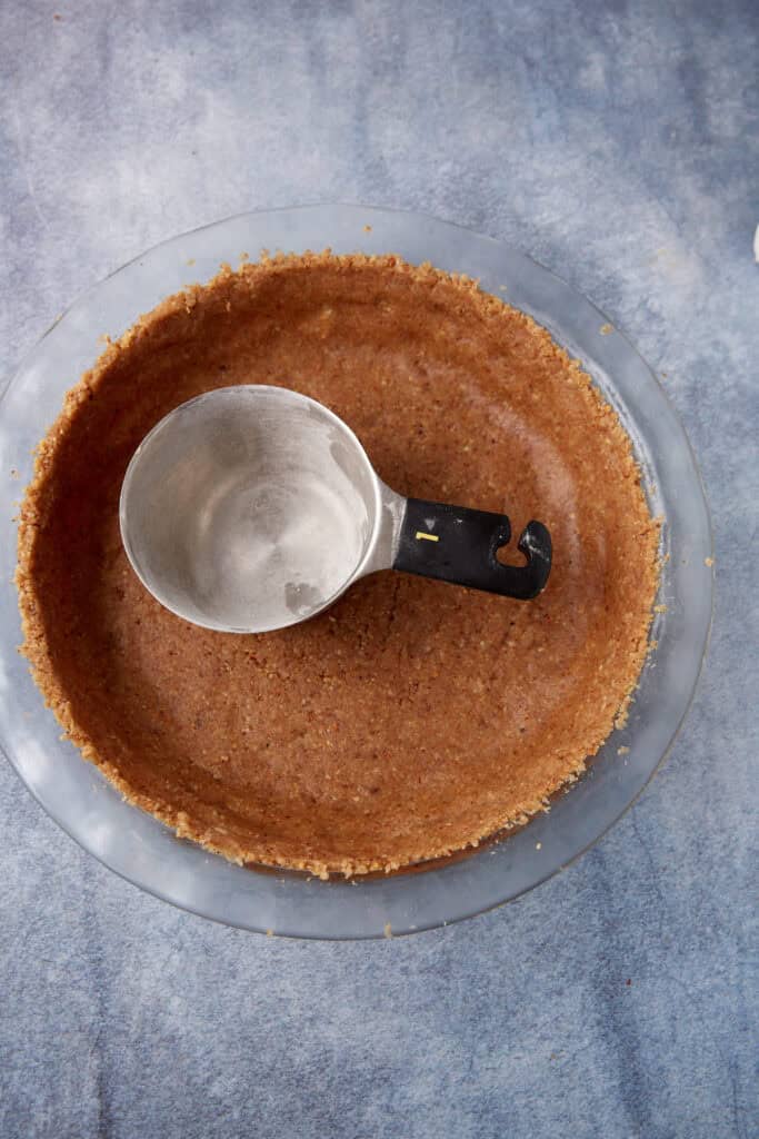 pecan crust in a glass pie plate flattened out with a measuring cup 