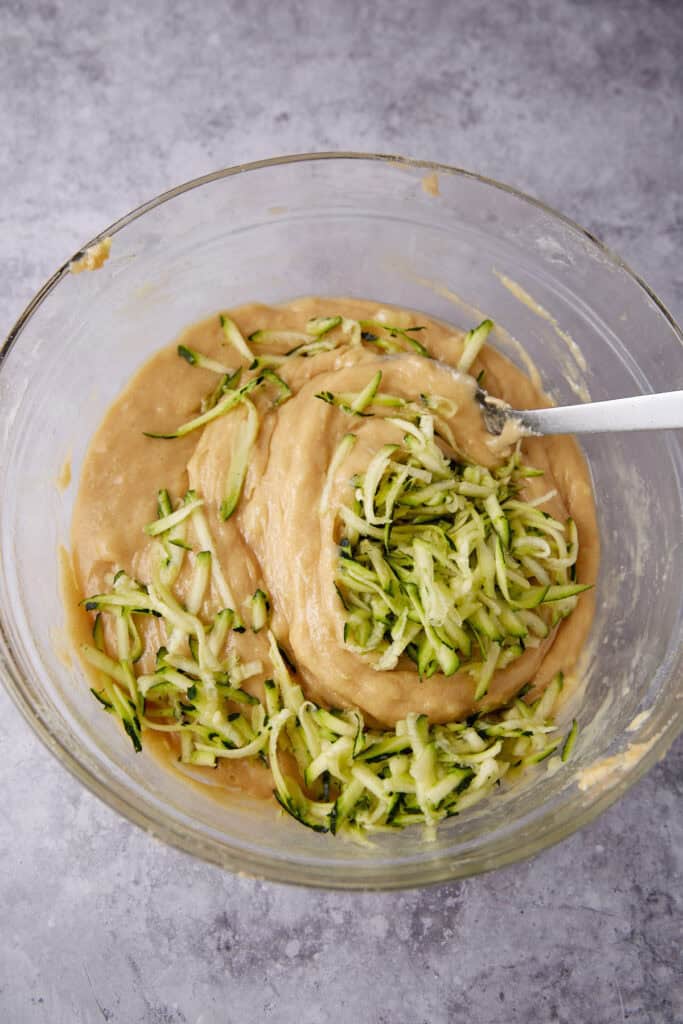 batter with zucchini being folded into it with a metal spoon in a glass bowl. 