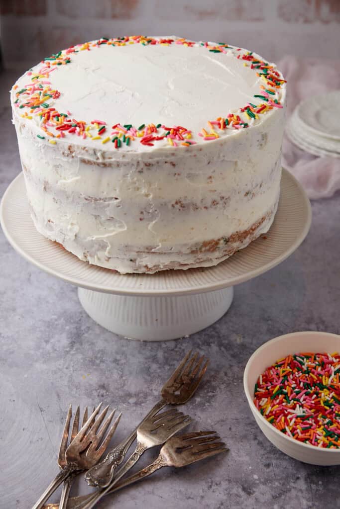 frosted confetti cake on a white cake stand with sprinkles on top.