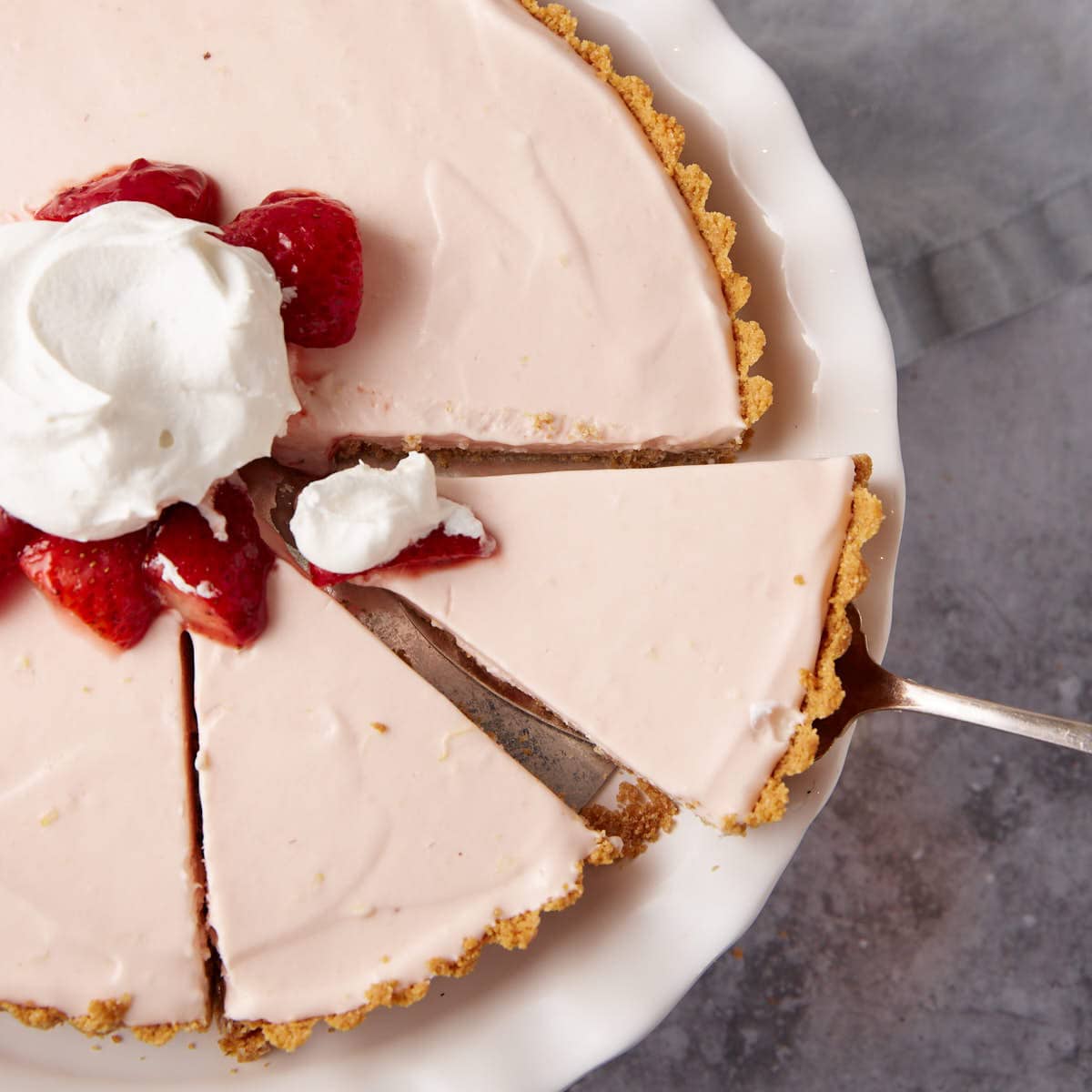 frozen strawberry pie topped with marinated strawberries and whipped cream on a white cake stand with a slice being lifted out.