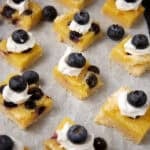 lemon blueberry bars on parchment paper topped with a dollop of whipped cream and a blueberry on top.