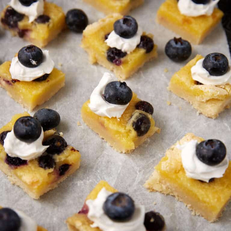 lemon blueberry bars on parchment paper topped with a dollop of whipped cream and a blueberry on top.