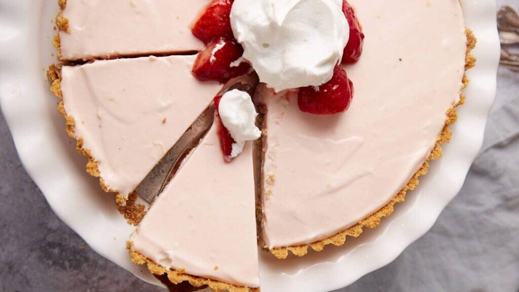 frozen strawberry pie topped with marinated strawberries and whipped cream on a white cake stand with a slice being lifted out.