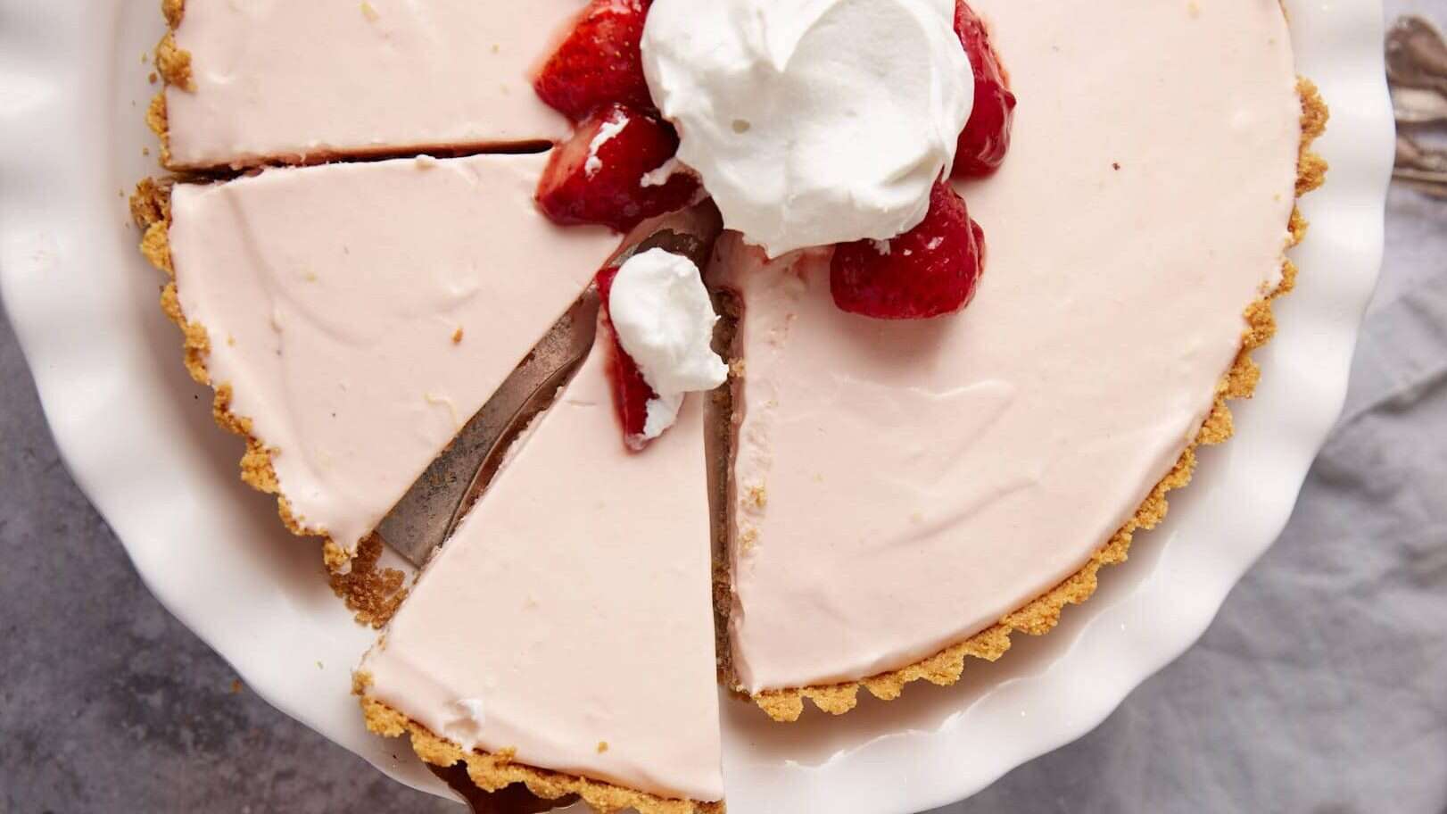 frozen strawberry pie topped with marinated strawberries and whipped cream on a white cake stand with a slice being lifted out.