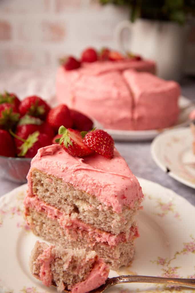 slice of strawberry cake with a bite taken out of it on a decorate plate with the strawberry cake in the background.