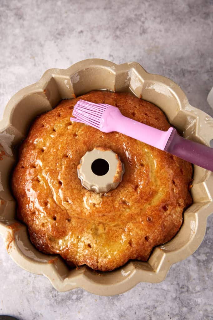 A cake in a pan being brushed with a pastry brush.