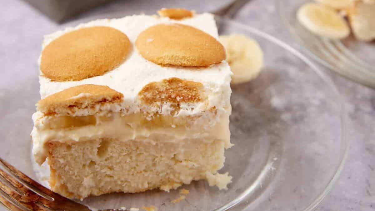 slice of banana pudding poke cake on a glass plate with a fork on the side and the cake in the background.