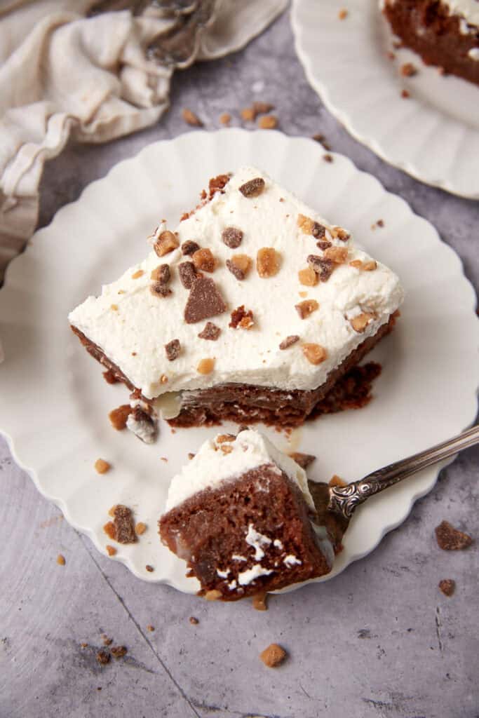 piece of heath bar cake on a white plate with a fork taking a bite out of it.
