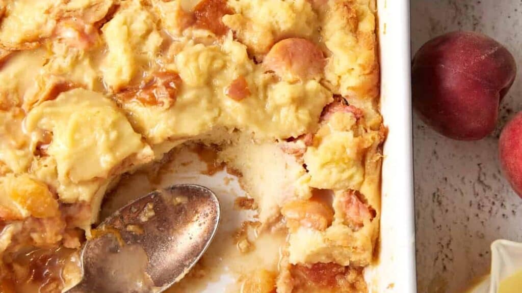 A casserole dish with peach bread pudding with part of it removed.