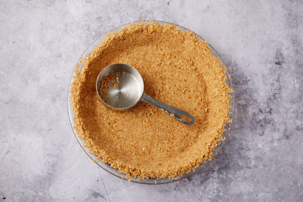 pretzel crust being pressed into a pie dish with a measuring cup. 