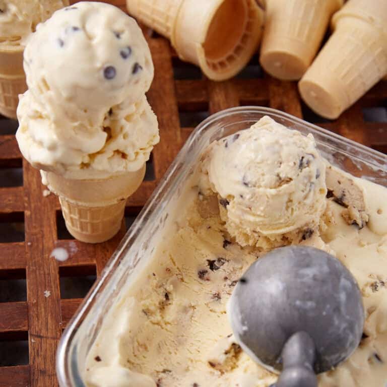 chocolate chip cookie dough ice cream being scooped out of a glass container with a cone and two scoops of ice cream on it.