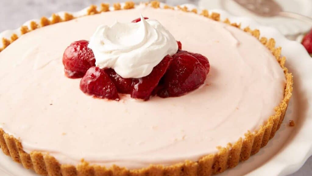 frozen strawberry pie topped with marinated strawberries and whipped cream on a white cake stand.