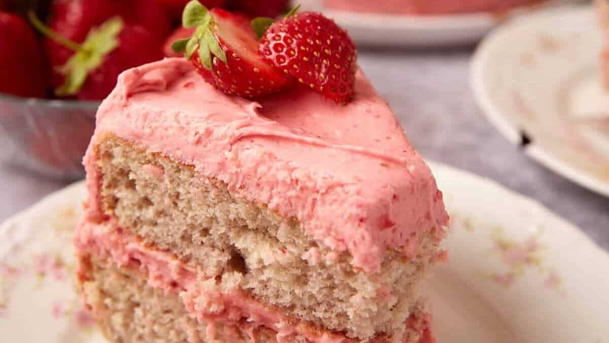 slice of strawberry cake with a bite taken out of it on a decorate plate with the strawberry cake in the background.