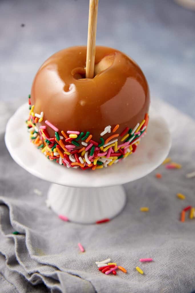caramel apple dipped in rainbow sprinkles on a white cake stand.