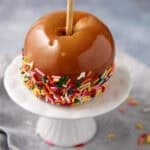 caramel apple dipped in rainbow sprinkles on a white cake stand.