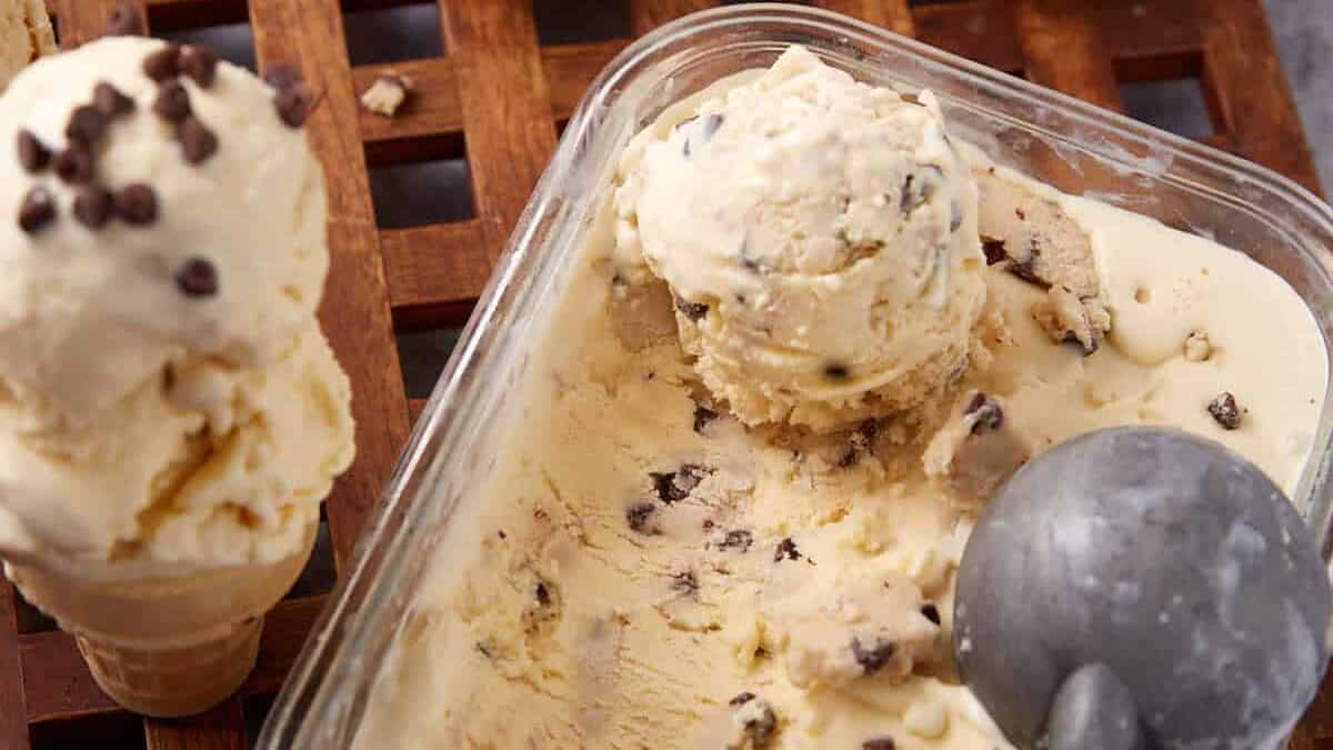 chocolate chip cookie dough ice cream being scooped out of a glass container with a cone and two scoops of ice cream on it.
