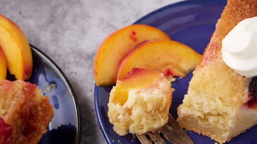 a piece of peach upside down cake on a blue plate with a fork cutting into it.
