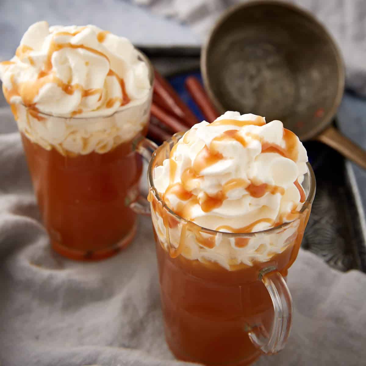 2 clear mugs filled with slow cooker apple cider topped with whipped cream and caramel drizzle.