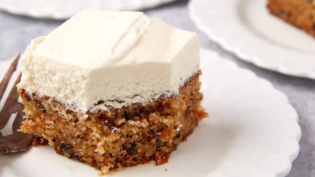 close up of a slice of carrot cake sheet cake on a white plate with a fork and a bite taken out of it.