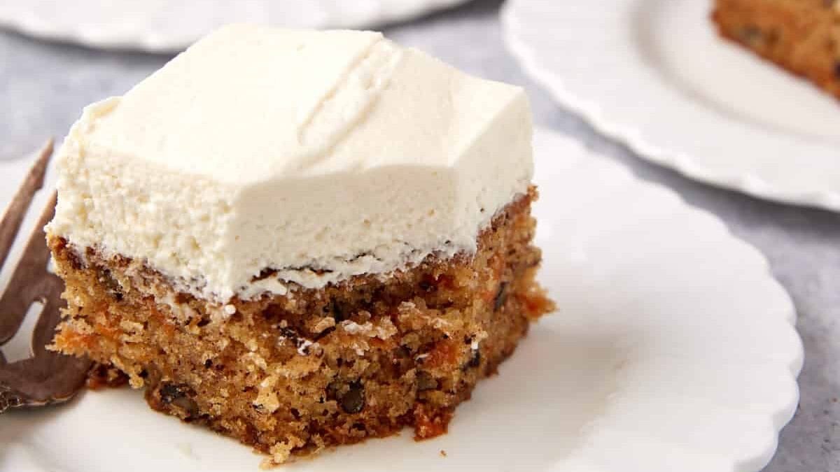 close up of a slice of carrot cake sheet cake on a white plate with a fork and a bite taken out of it.