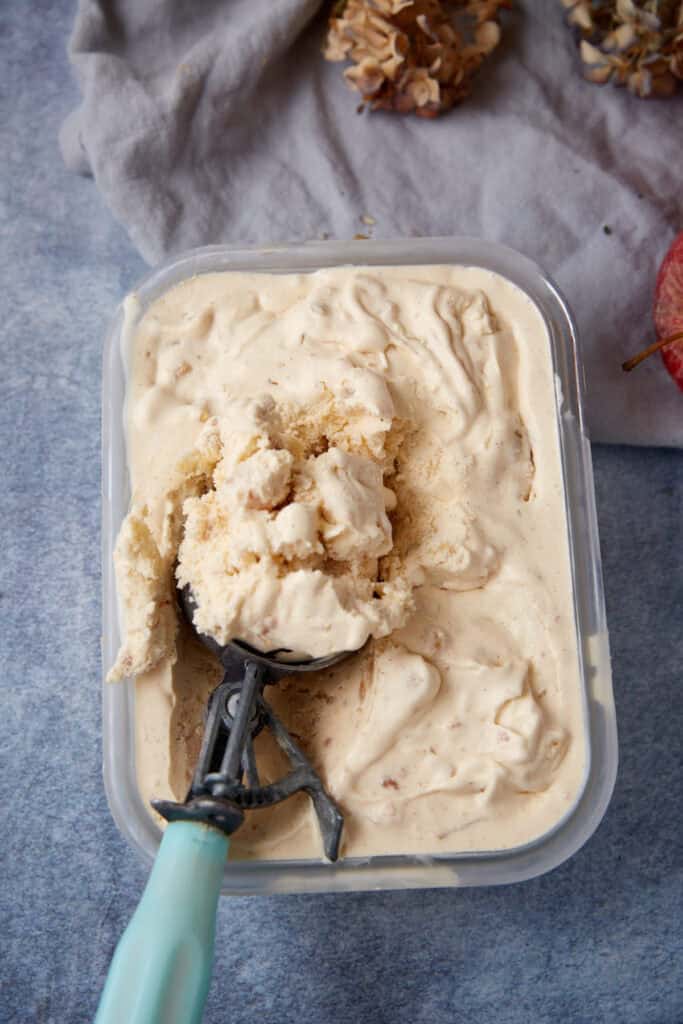 Homemade apple crisp ice cream being scooped out of a rectangular container with a vintage blue-handled scoop.