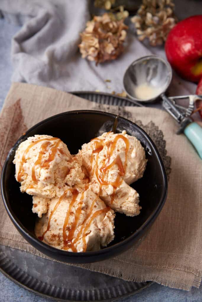 Overhead view of apple crisp ice cream scooped into a black bowl, set on a tan linen with an ice cream scoop and dried flowers in the background.