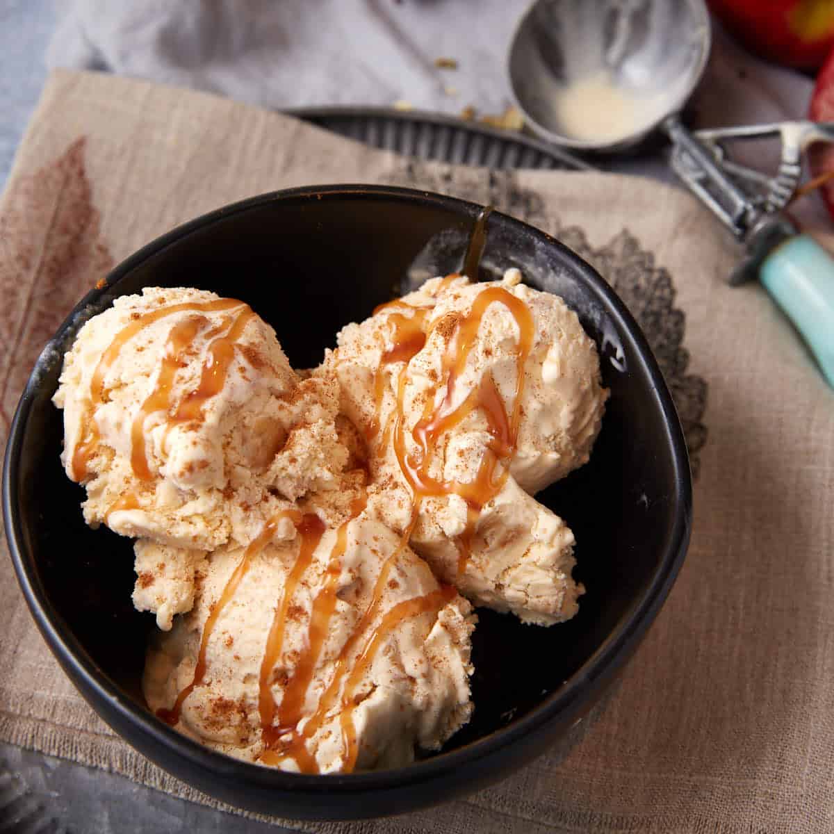Overhead shot of a bowl of apple crisp ice cream with caramel drizzle, set on a linen napkin with an ice cream scoop and apples in the background.