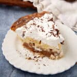 A slice of Banoffee Pie on a white plate with layers of caramel, bananas, and whipped cream topped with chocolate shavings.