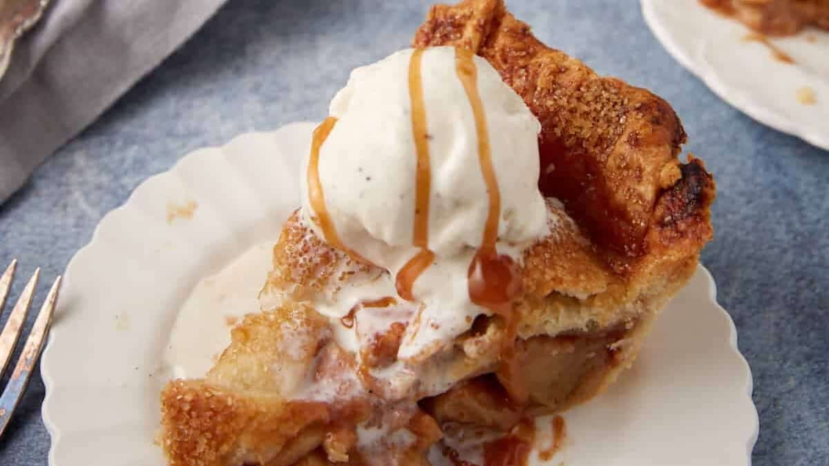 slice of caramel apple pie on a white plate with a scoop of vanilla ice cream and a drizzle of caramel.