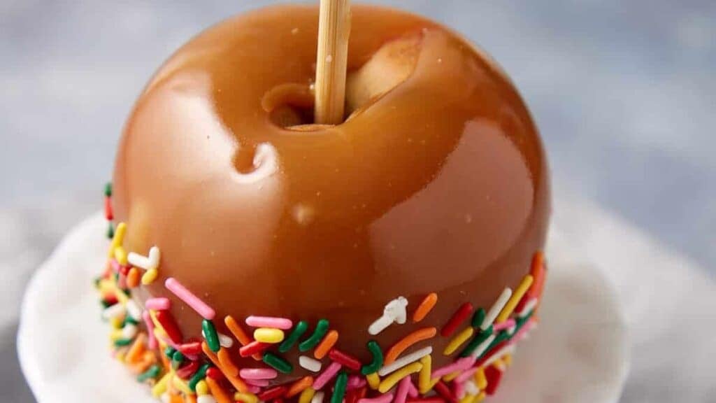 caramel apple dipped in rainbow sprinkles on a white cake stand.