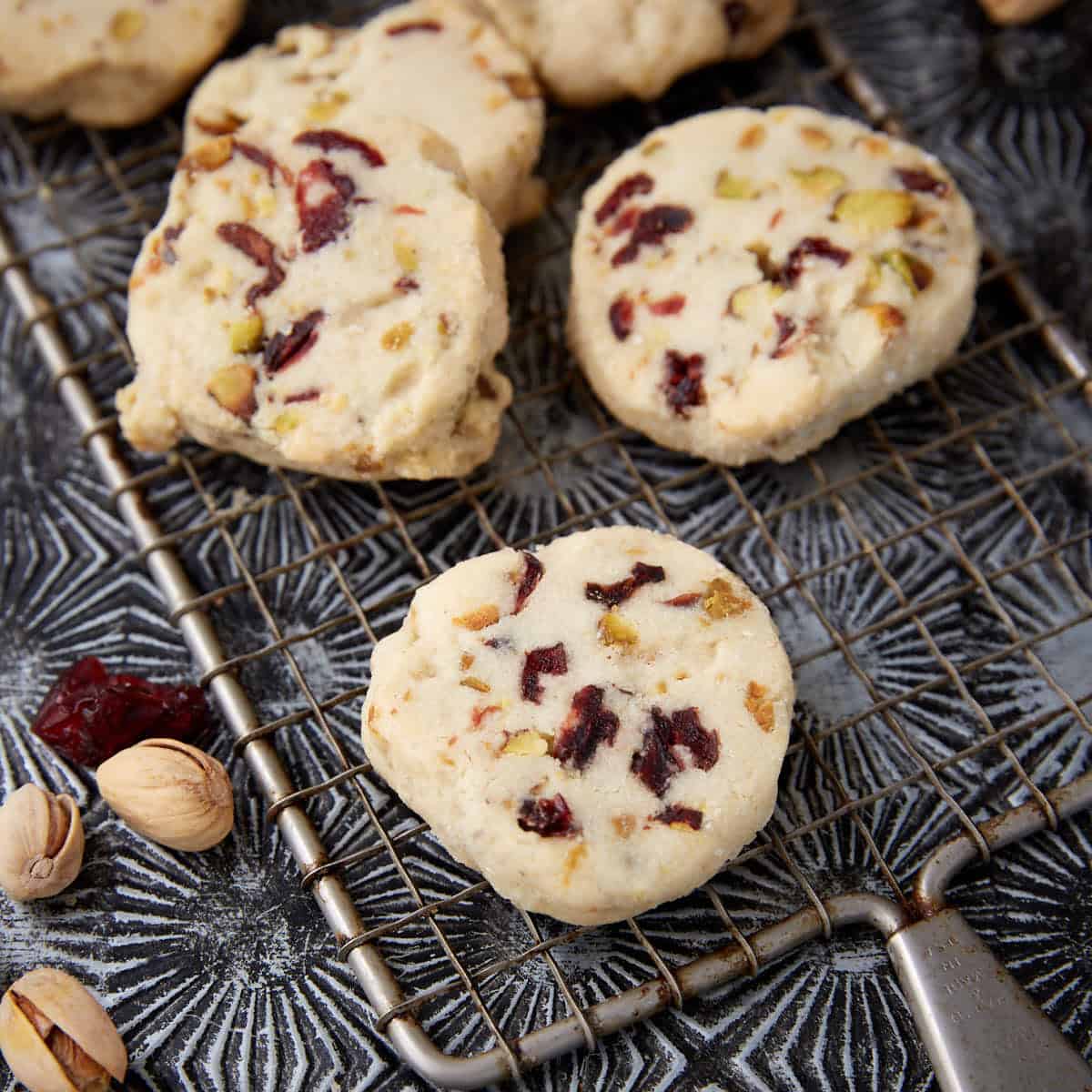 Close-up of cranberry pistachio shortbread cookies on a cooling rack, with chopped pistachios and dried cranberries scattered nearby.