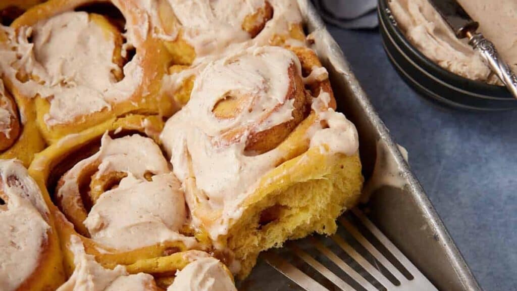 iced pumpkin cinnamon rolls in a baking dish with a spatula.