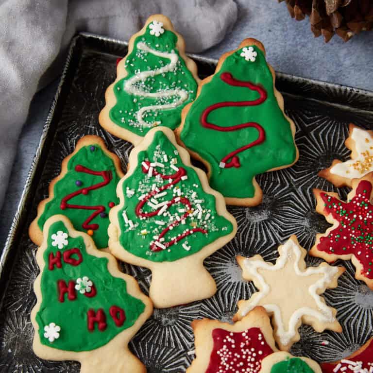 Close-up view of decorated Christmas tree and star sugar cookies on a baking tray, with a pine cone and a cloth in the background.
