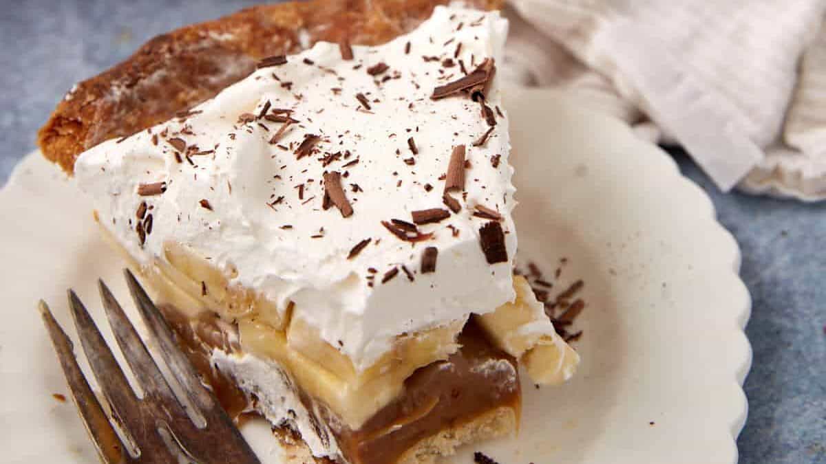 A plated slice of Banoffee Pie with a fork beside it, showing caramel and banana layers under whipped cream.