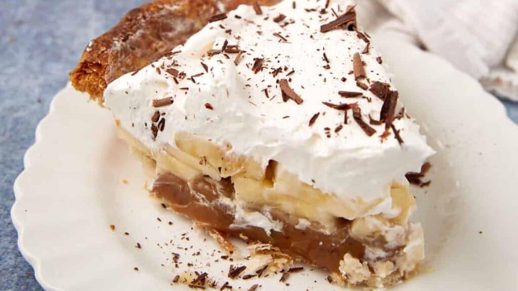 A slice of Banoffee Pie on a white plate with layers of caramel, bananas, and whipped cream topped with chocolate shavings.