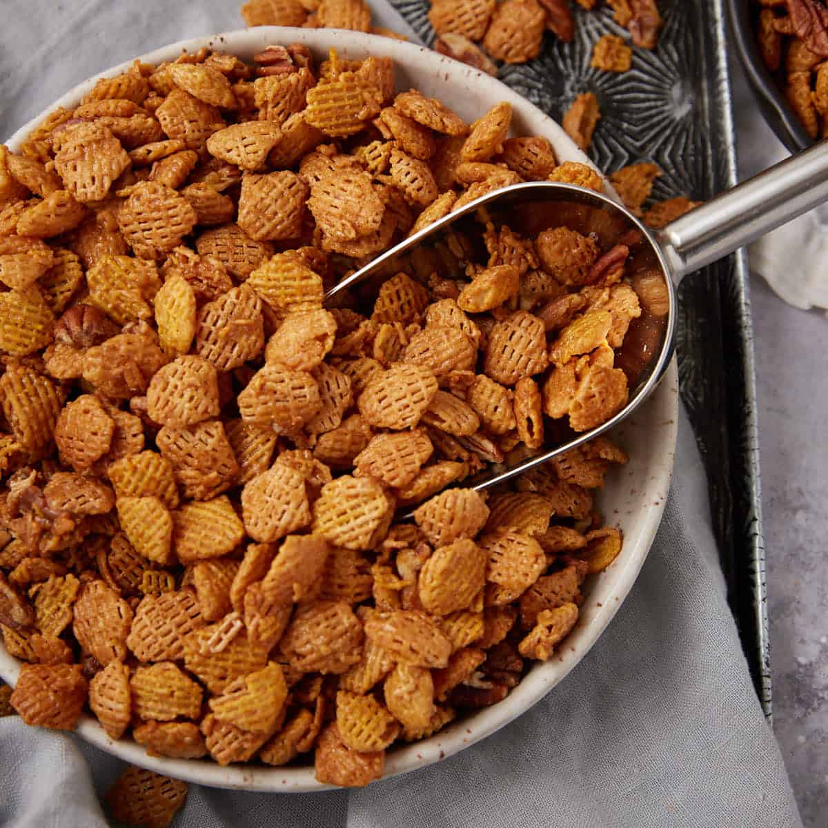 Close-up of a serving scoop resting in a large bowl of caramel cereal mix, showing the glossy coating.