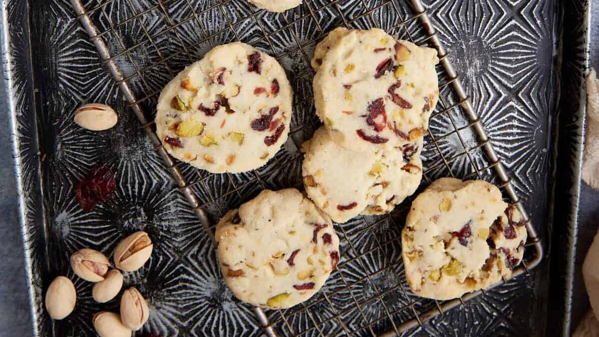 Overhead view of several cranberry pistachio shortbread cookies on a cooling rack and baking tray, surrounded by shelled pistachios and dried cranberries.