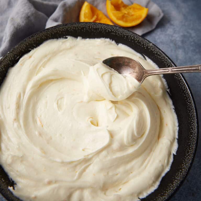 A close-up of a black bowl of orange cream cheese frosting, with a spoon resting on top and visible flecks of orange zest throughout the frosting.