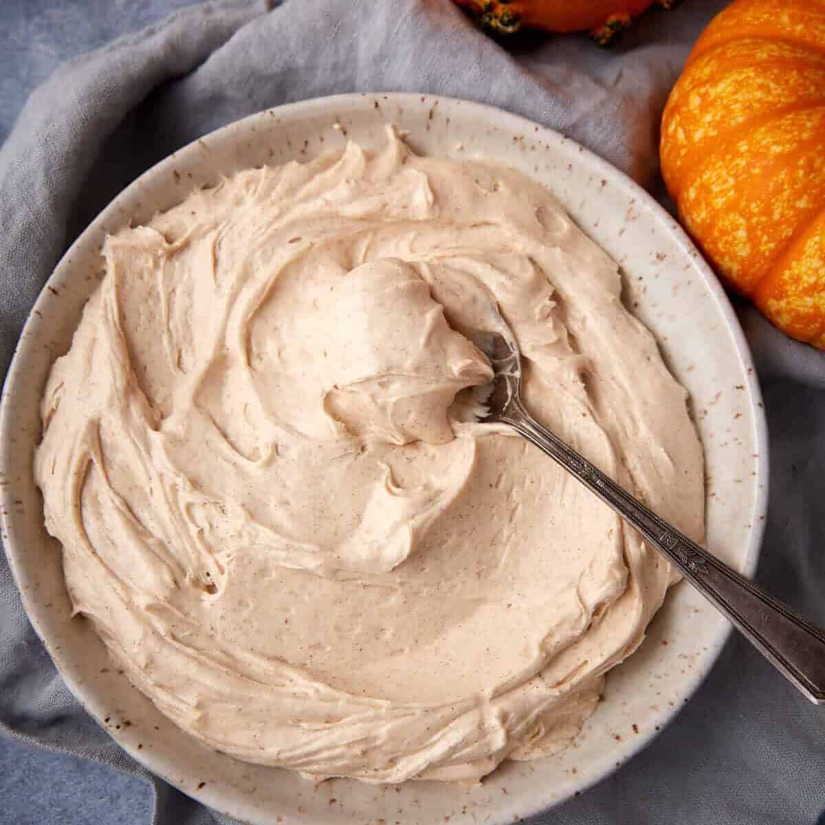A bowl of finished pumpkin spice cream cheese frosting with a spoon, surrounded by small pumpkins and a linen cloth.