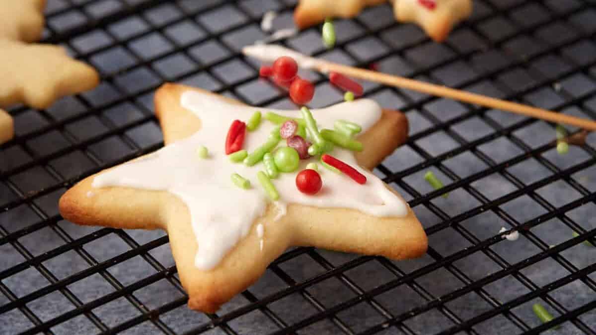 A hand sprinkling red, green, and white holiday sprinkles over a star-shaped sugar cookie with white royal icing on a cooling rack.