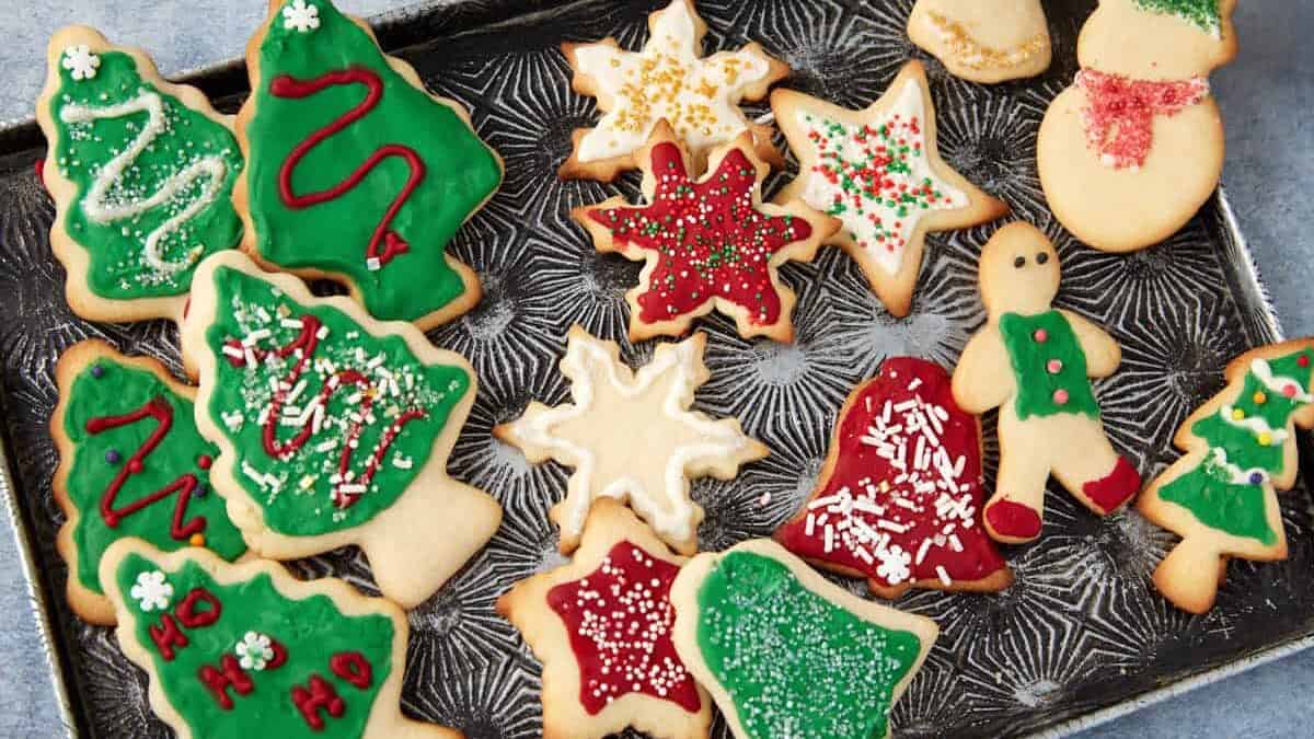 A baking tray filled with decorated holiday sugar cookies, including Christmas trees, snowflakes, stars, a bell, a gingerbread person, and a snowman, all with colorful icing and sprinkles.