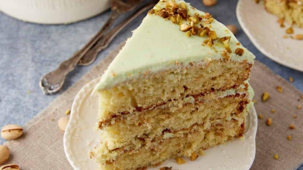 Slice of Watergate Cake on a white plate, showing pale green frosting and three pistachio-studded layers.