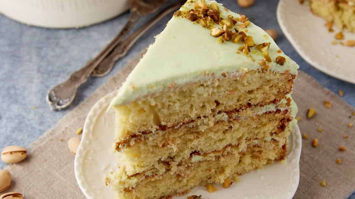 Slice of Watergate Cake on a white plate, showing pale green frosting and three pistachio-studded layers.