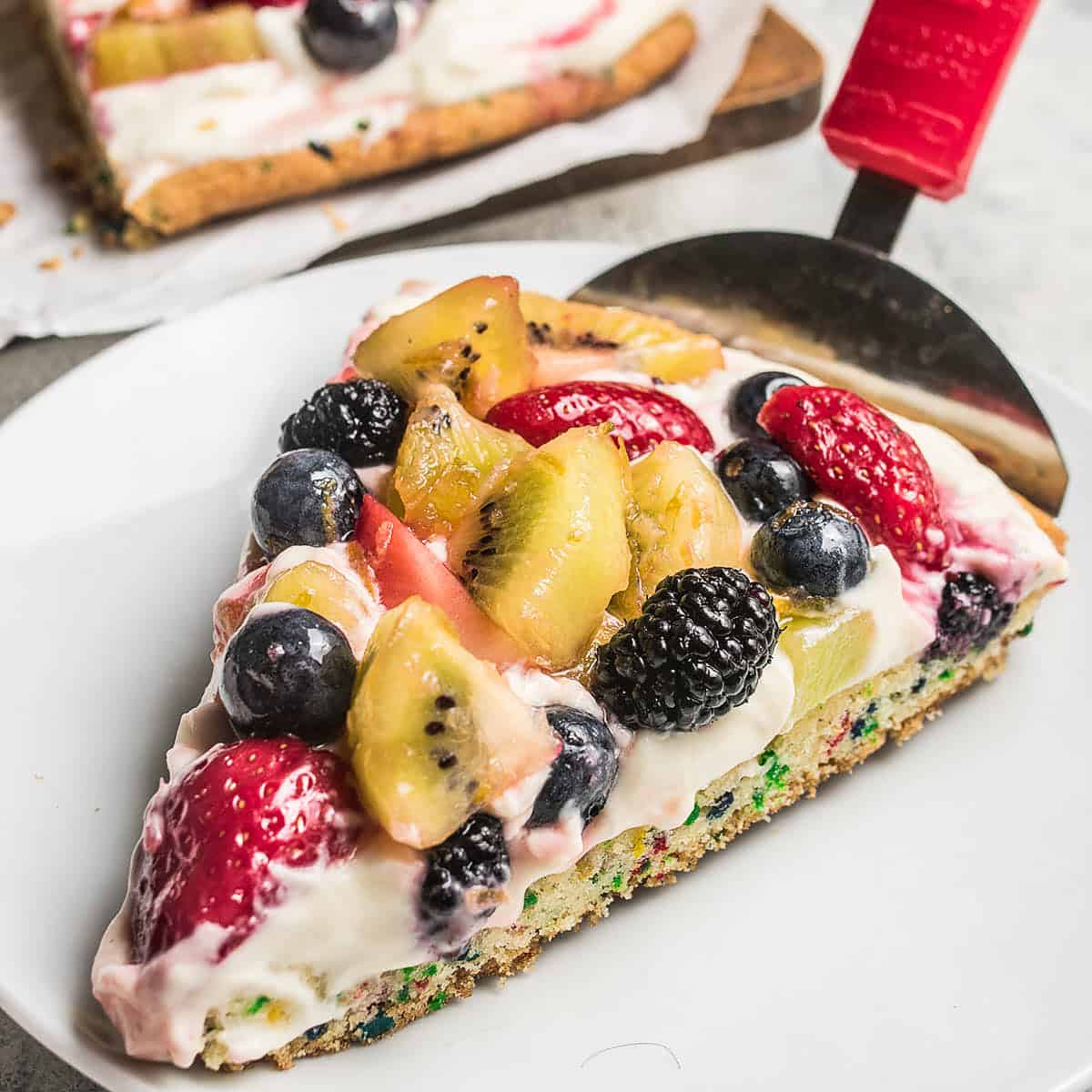 Slice of fruit pizza on a white plate topped with strawberries, kiwi, blueberries, and blackberries over cream cheese frosting.