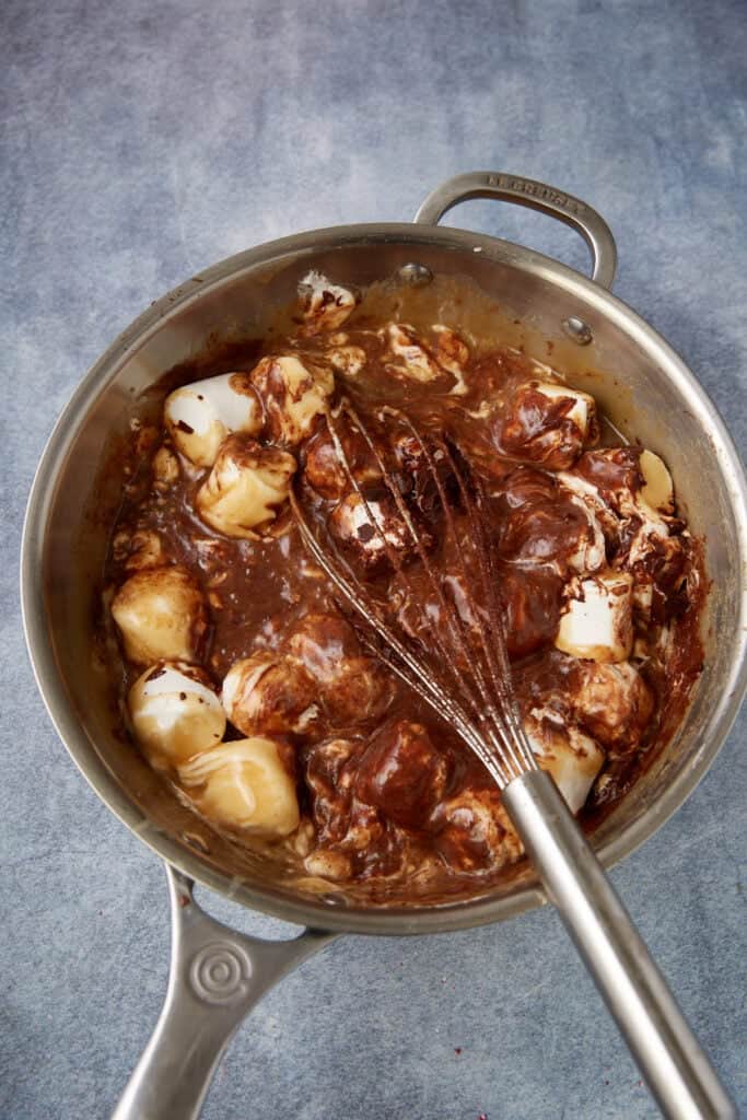 Marshmallows melting into warm chocolate mixture in a saucepan while being whisked for peppermint fudge.