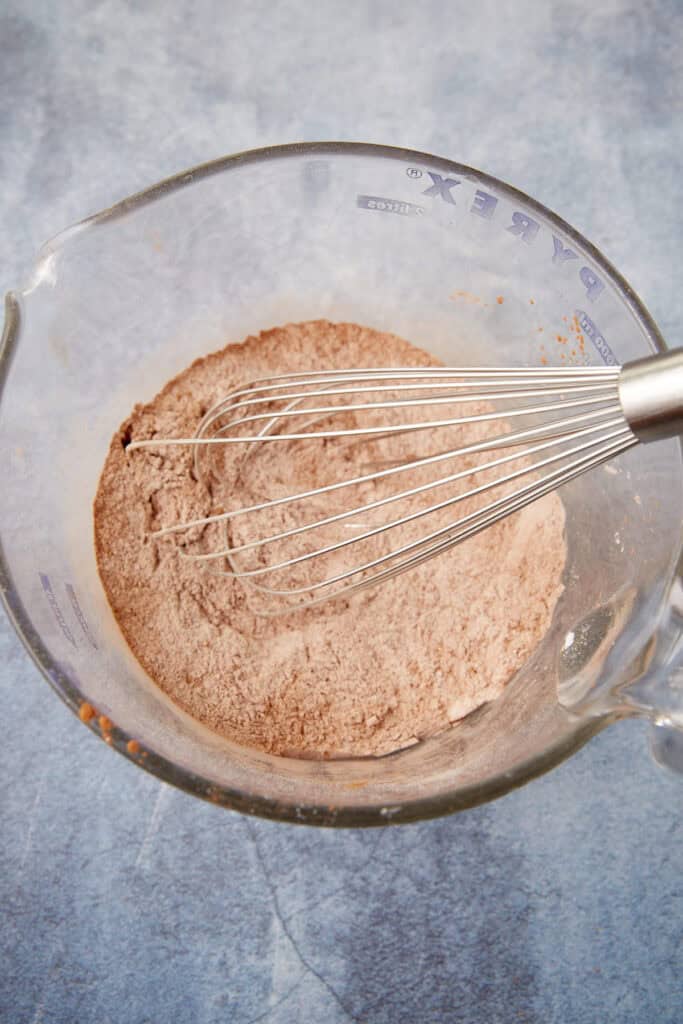 Glass measuring bowl filled with whisked dry chocolate cake ingredients and a metal whisk resting on top.