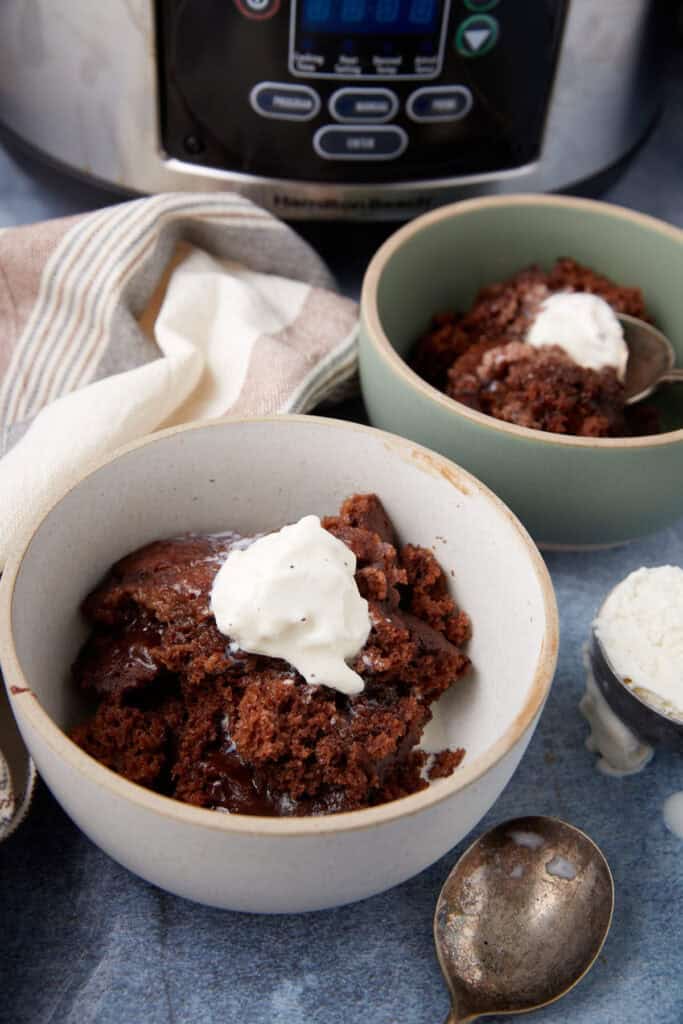 Two bowls of slow cooker chocolate lava cake with ice cream in front of a slow cooker, with a spoon and a small scoop of extra ice cream on the side.